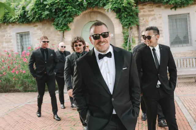 Anthony in a black tuxedo with his wedding party outside the stone villa.